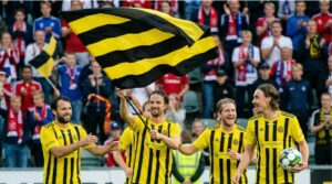 Spelare i svartgula matchtr&ouml;jor firar seger framf&ouml;r fullsatta l&auml;ktare med en stor svartgul flagga, symbol f&ouml;r klubbens starka community.